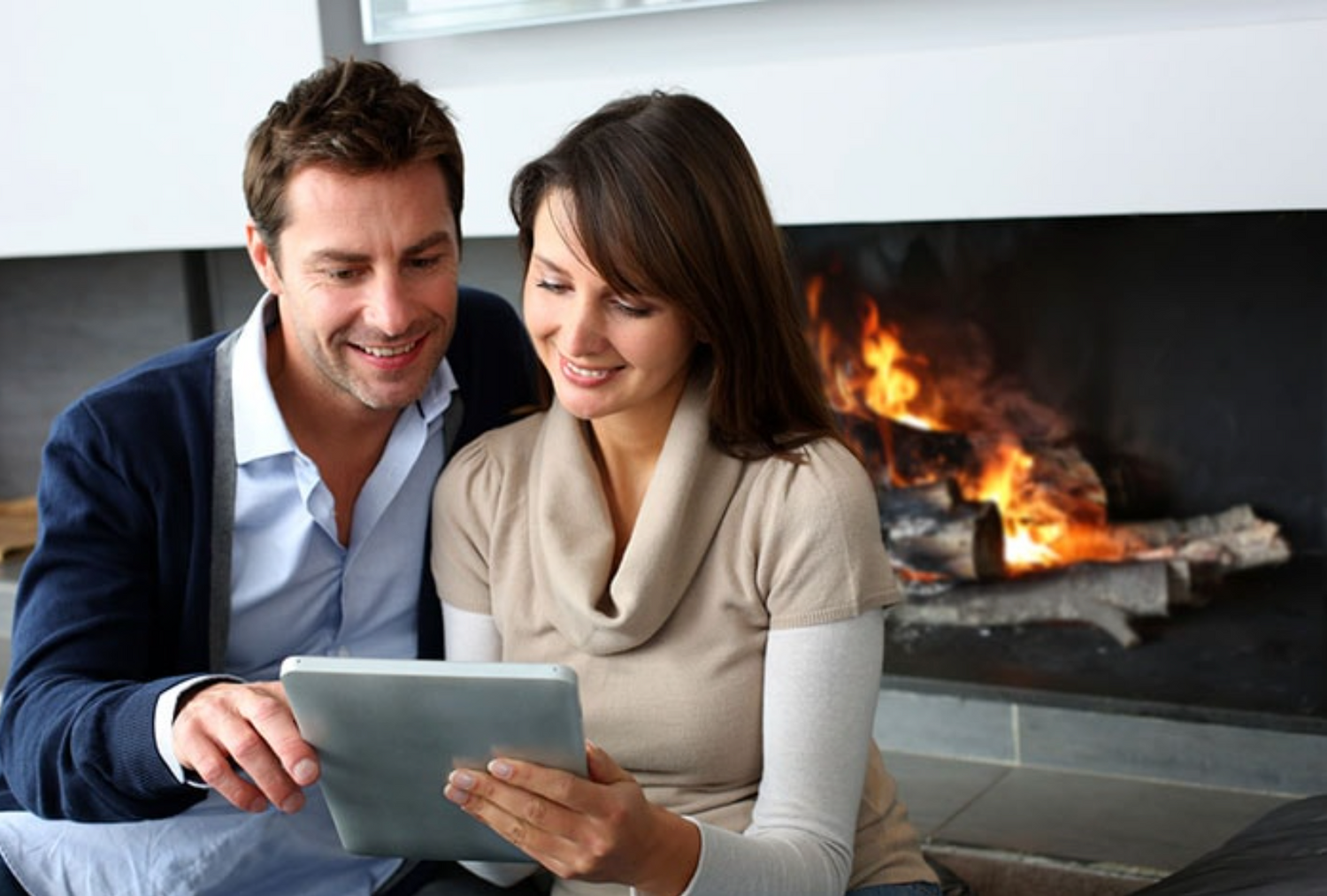 A man and woman use a tablet together in front of a fireplace
